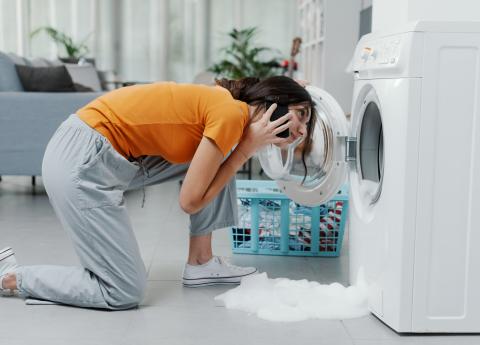 Woman kneeling on the floor holding a phone while looking into a broken washing machine