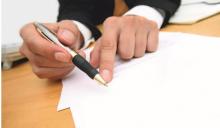 A man wearing a business suit and holding a pen, pointing at a piece of paper